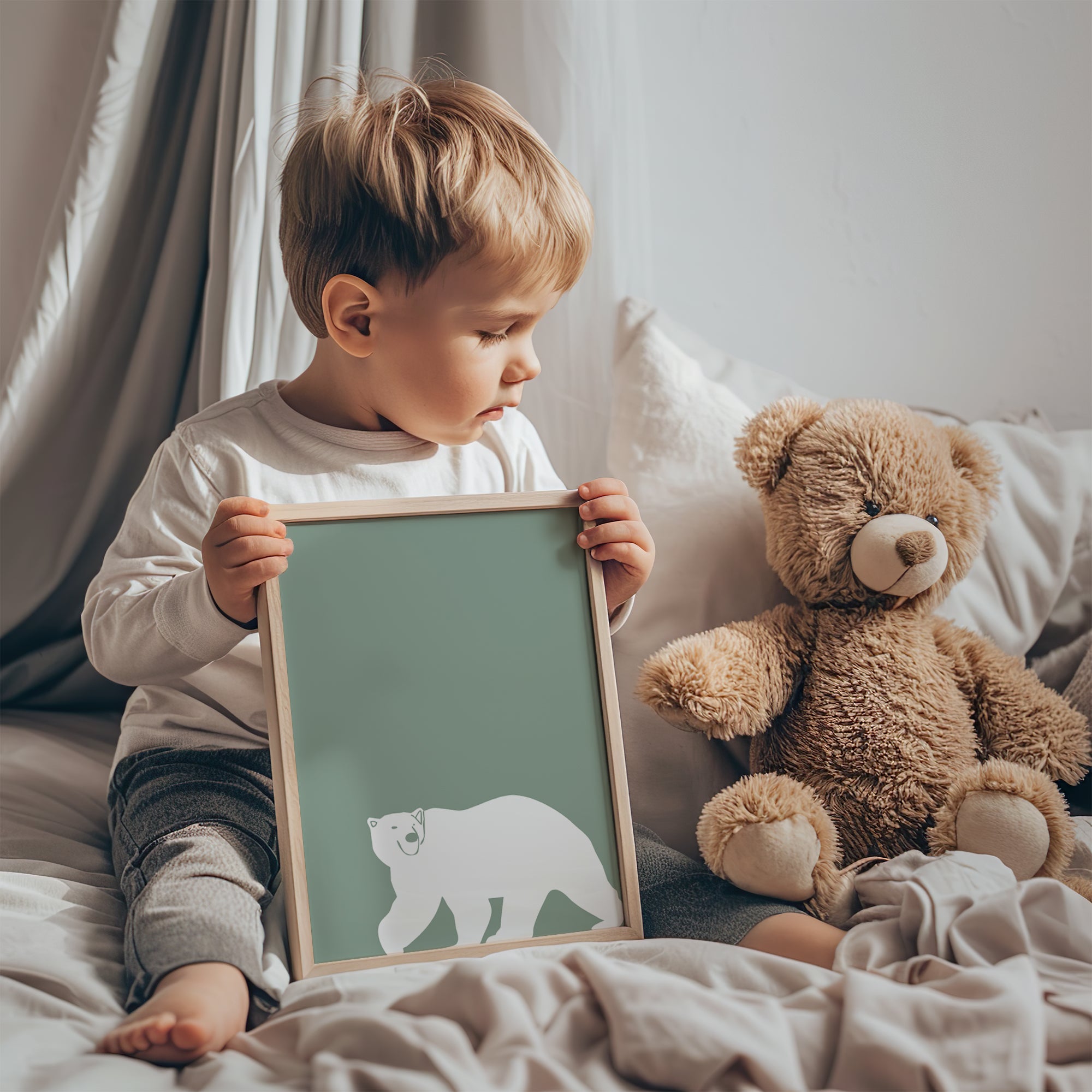young boy holding a framed green polar bear print while sitting beside a teddy bear on a bed