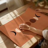 A terracotta-colored yoga mat with a minimalist face graphic, being held by hands on a light wooden table in warm natural sunlight. A 'The Hall Habitat' logo is visible in the bottom right corner.