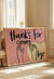 Framed pink wall art print with black handwritten text reading 'Thanks for coming, bye' and hand illustrations over a heart, leaning against the wall in a bright living room